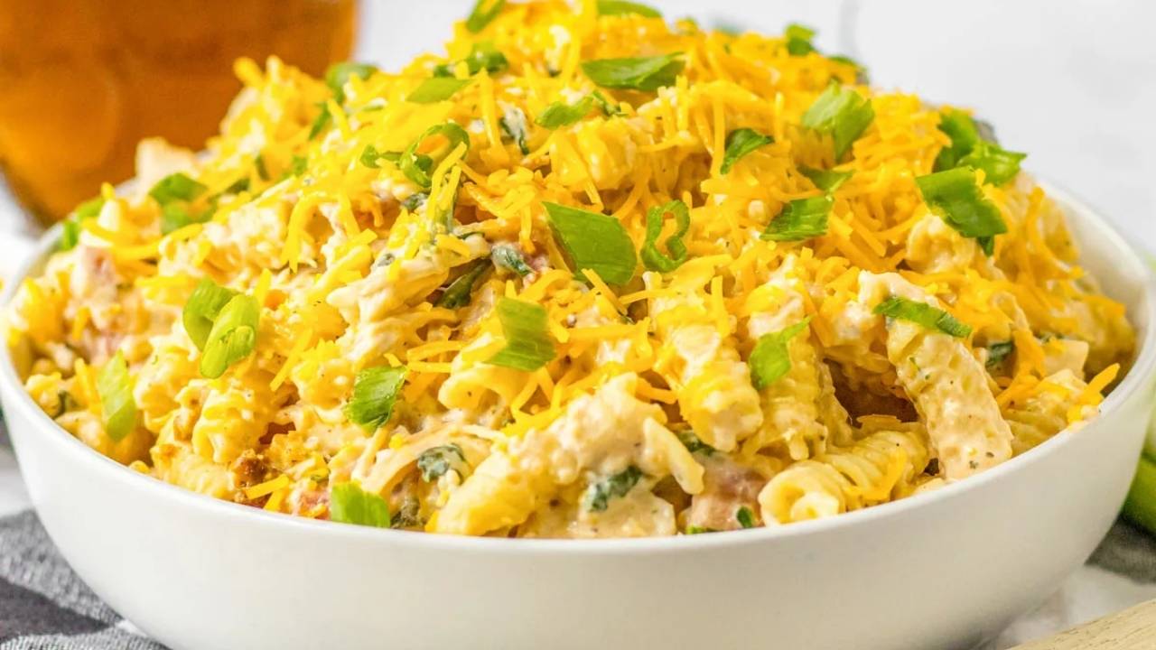 A bowl of creamy pasta salad topped with shredded cheddar cheese and chopped green onions.