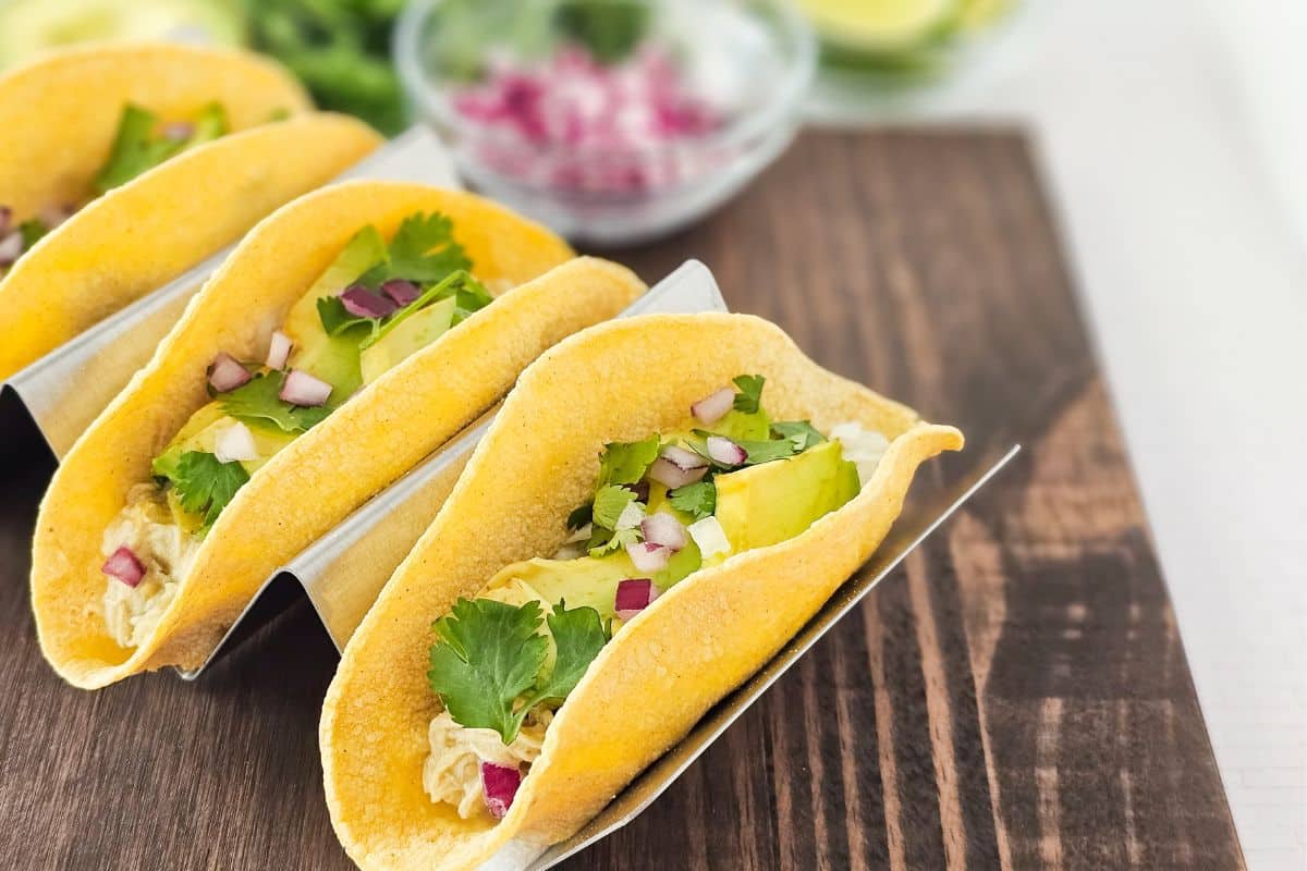 Three tacos filled with shredded chicken, avocado, cilantro, and diced red onion are arranged in a metal taco holder on a wooden surface.
