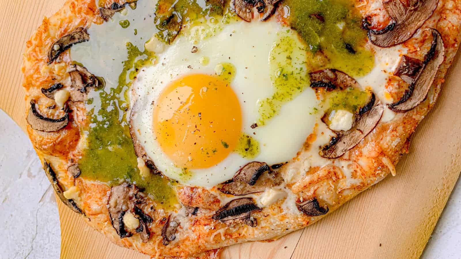 A baked flatbread topped with a sunny-side-up egg, sliced mushrooms, melted cheese, and a drizzle of green sauce.
