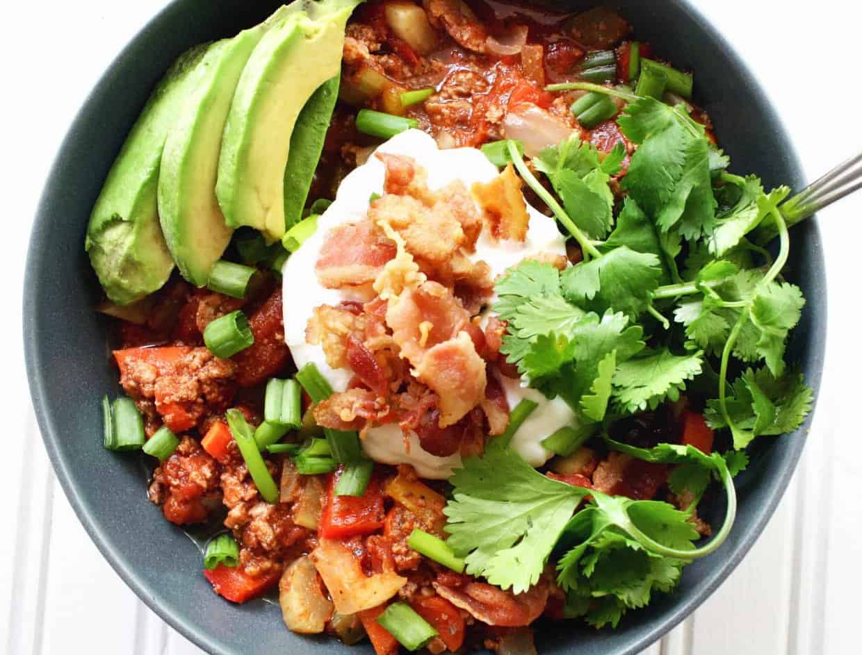 A bowl of chili topped with sliced avocado, sour cream, cooked bacon, chopped green onions, and fresh cilantro.