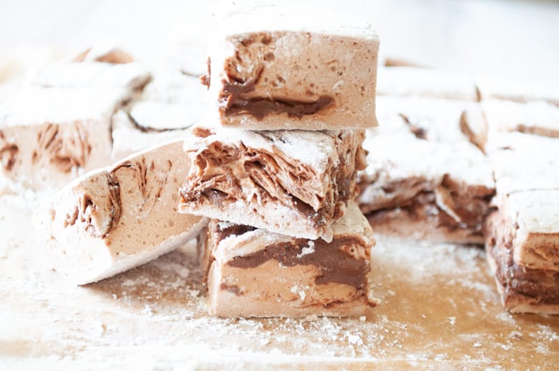 Several pieces of chocolate and vanilla marbled marshmallows dusted with powdered sugar are stacked on a wooden surface.