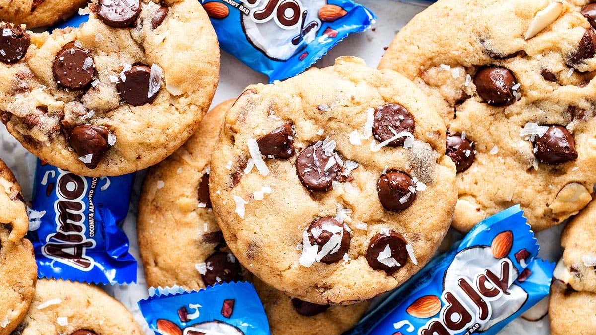 Chocolate chip cookies topped with sea salt are arranged on a surface alongside unwrapped blue Almond Joy mini candy bars.