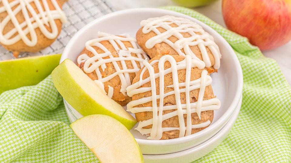 Three cookies with white icing drizzle are served in a white bowl with two slices of green apple, placed on a green checkered cloth. An apple and more cookies are in the background.