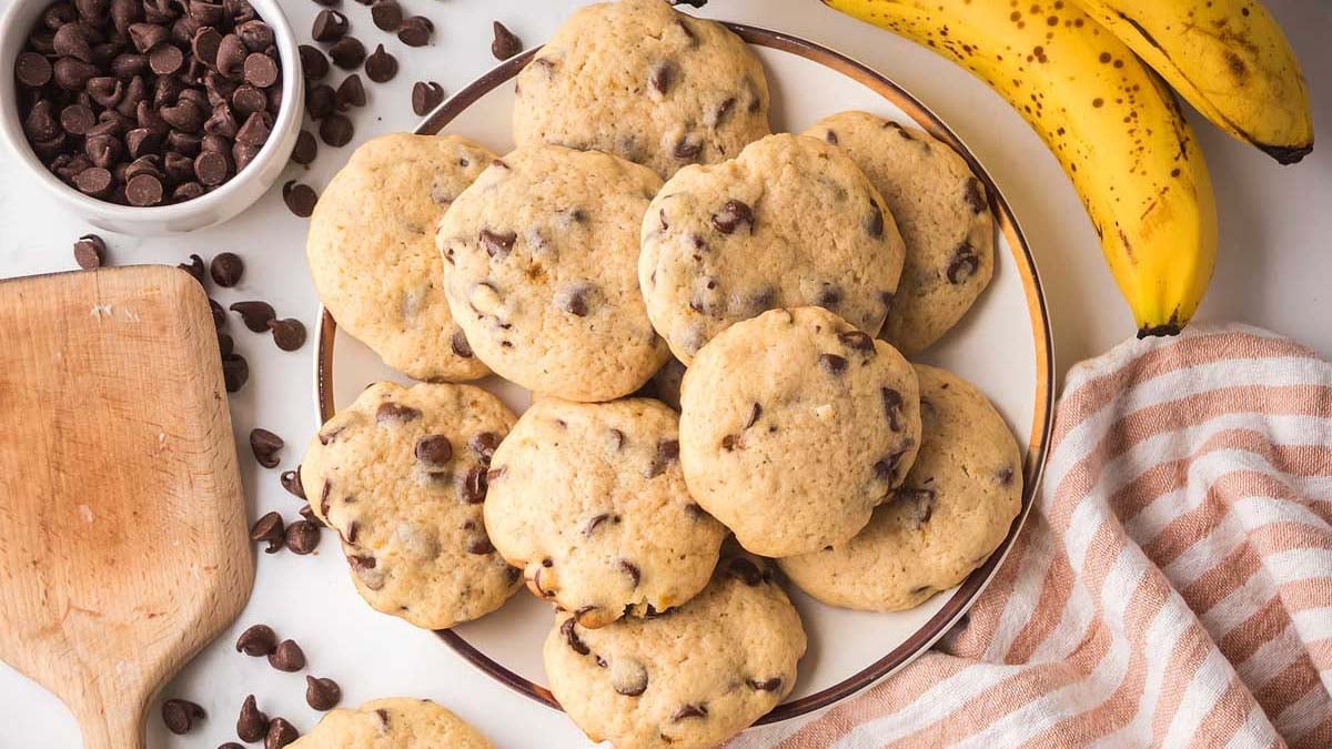 A plate of chocolate chip cookies is surrounded by bananas, a bowl of chocolate chips, a wooden board, and a striped cloth.