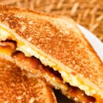 Close-up of a grilled sandwich with scrambled eggs, bacon, and melted cheese, placed on a white plate with a woven surface in the background.