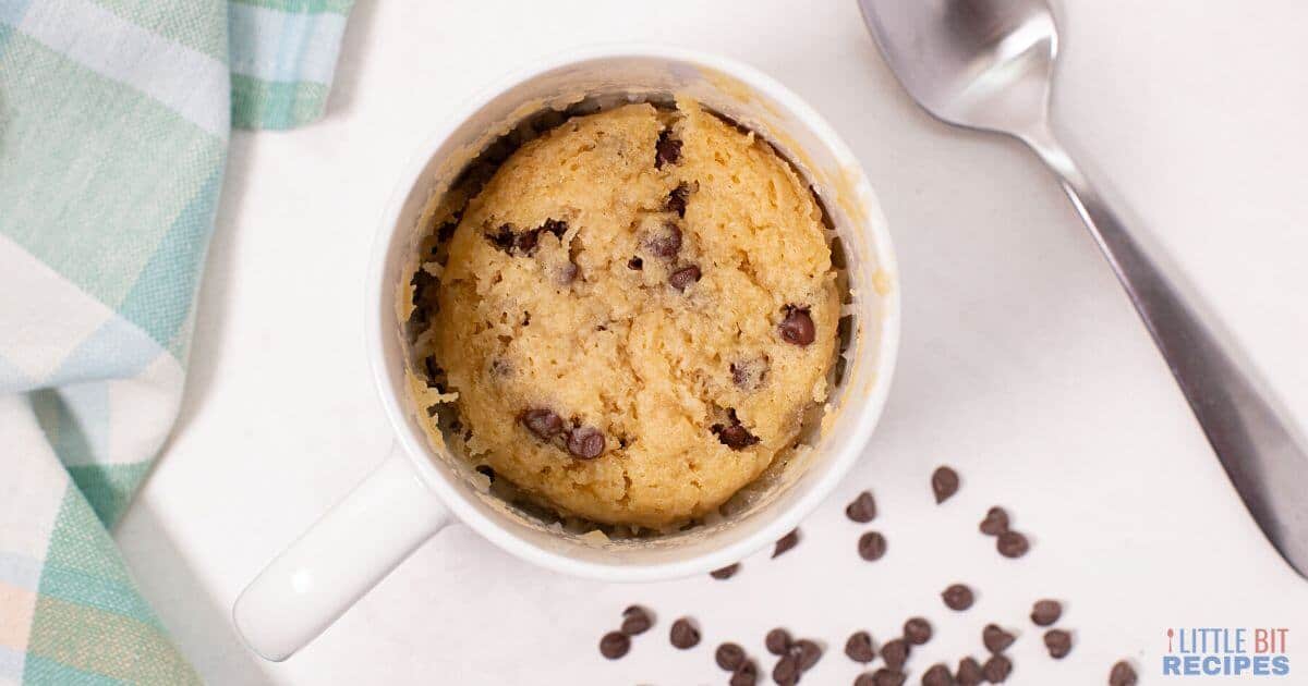 A chocolate chip cookie baked in a white mug sits on a white surface, with a spoon, chocolate chips, and a striped napkin nearby.