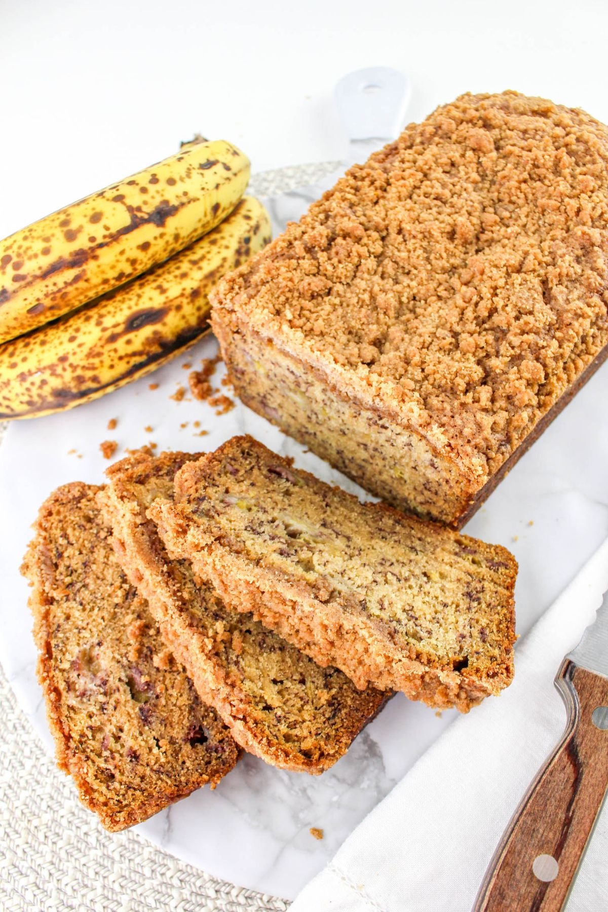 A loaf of banana bread with a crumb topping is partially sliced on a marble surface, next to a bunch of ripe bananas and a knife.