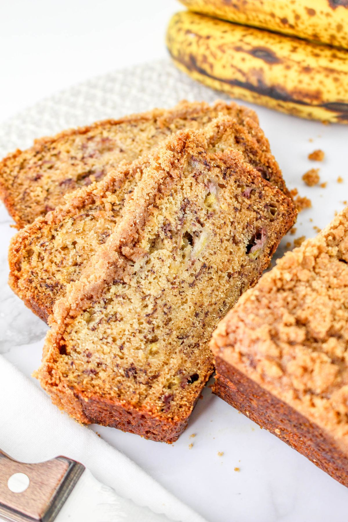 Slices of banana bread with a crumb topping are arranged on a marble surface, with ripe bananas in the background.