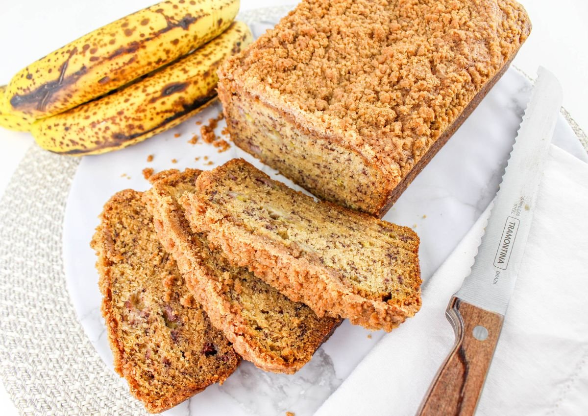 A loaf of banana bread with a crumb topping is partially sliced on a marble board next to ripe bananas and a serrated knife with a wooden handle.