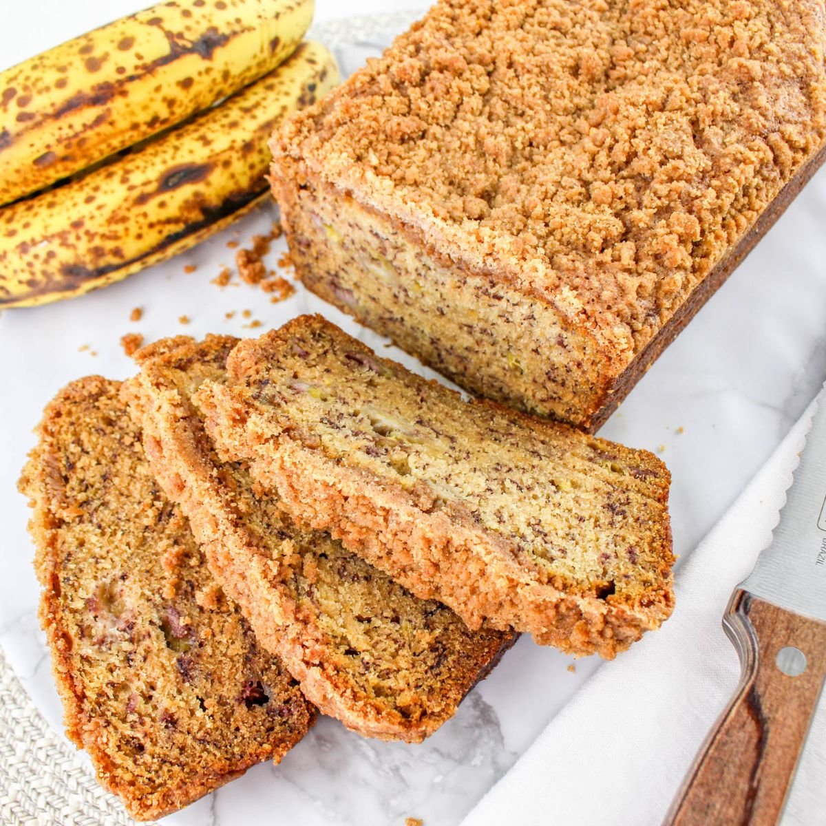 A loaf of banana bread with a crumb topping is sliced on a white surface, with ripe bananas and a knife nearby.