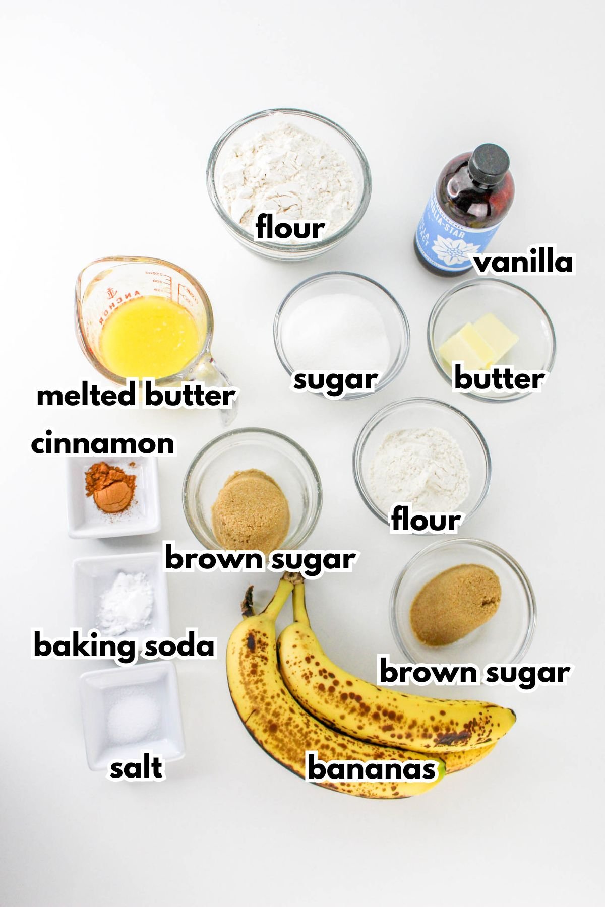 Overhead view of measured ingredients for banana bread, including flour, sugar, brown sugar, melted butter, butter, vanilla, cinnamon, baking soda, salt, and ripe bananas.
