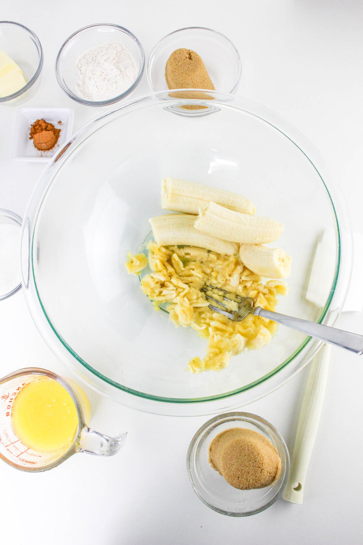 A glass bowl with mashed bananas and a fork, surrounded by measured ingredients including flour, sugar, butter, cinnamon, and brown sugar on a white surface.