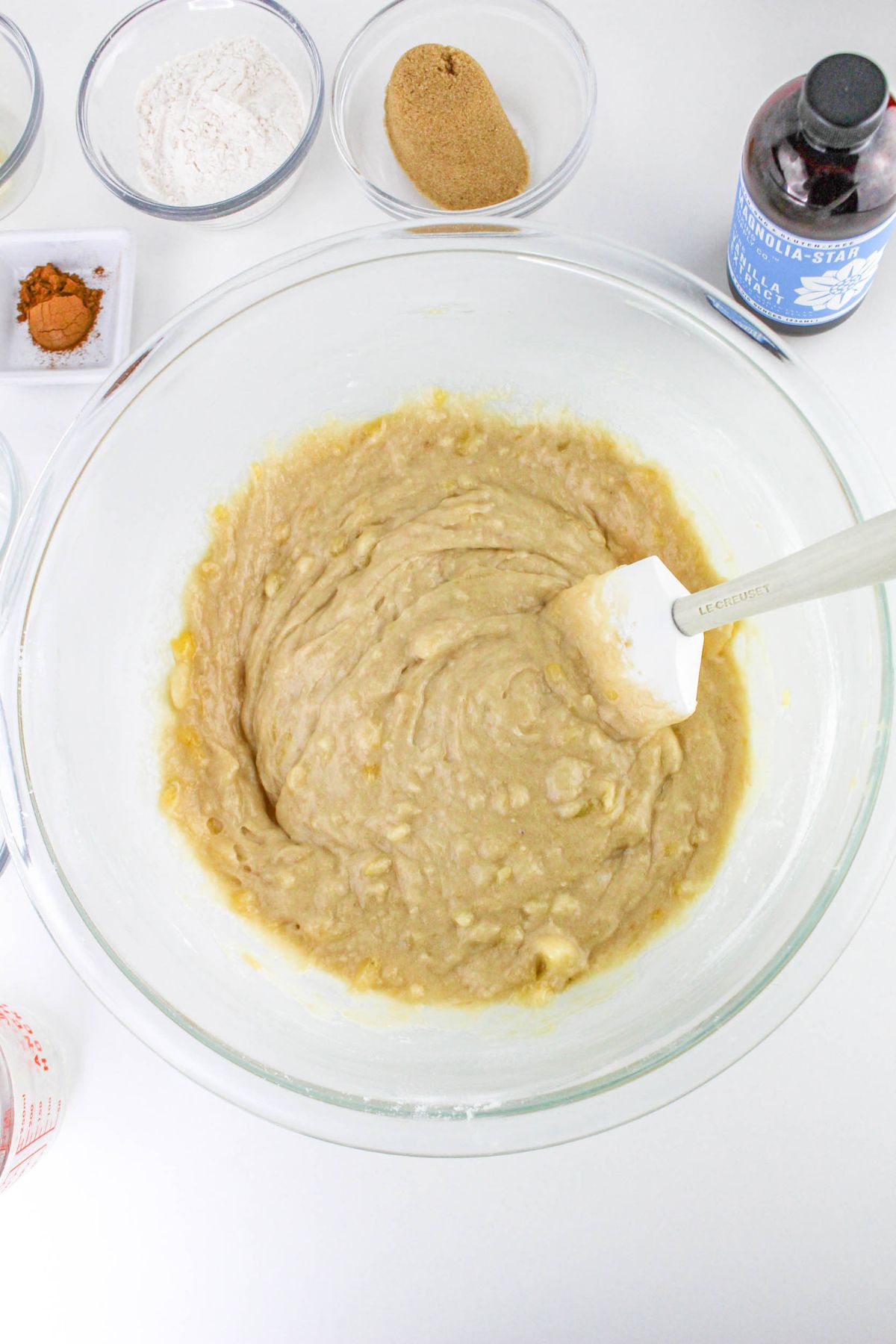 A glass bowl with banana bread batter being mixed with a spatula, surrounded by ingredients like flour, brown sugar, cinnamon, and vanilla extract on a white surface.