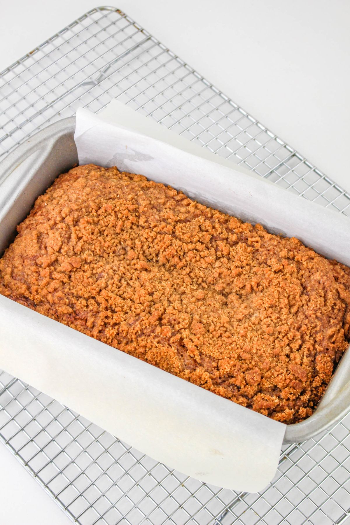 A loaf of crumb-topped bread in a parchment-lined baking pan, resting on a wire cooling rack.