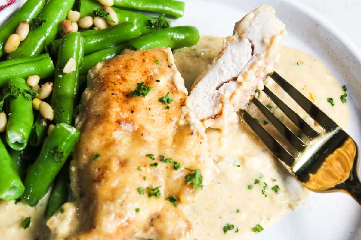 Sliced chicken breast in creamy sauce garnished with parsley, served with green beans and pine nuts on a white plate.