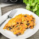 A serving of enchilada casserole with black beans, corn, and shredded chicken on a white plate with a fork, garnished with cilantro.
