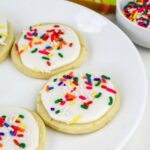 Three sugar cookies with white icing and colorful sprinkles on a white plate, with a bowl of sprinkles in the background.
