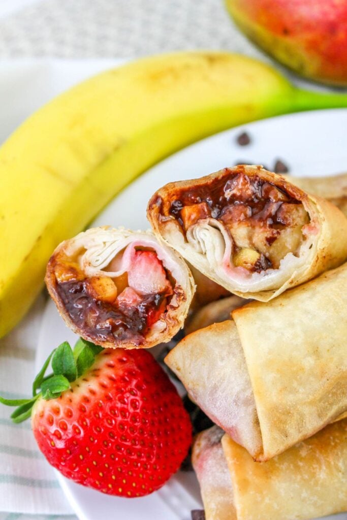 A plate with fruit-filled spring rolls, one cut open to show chocolate, banana, and strawberry inside, with a whole banana and a strawberry beside them.