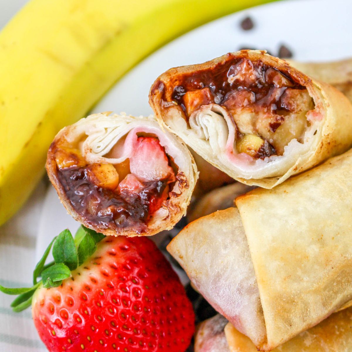 A close-up of sliced dessert spring rolls filled with bananas, strawberries, and chocolate, with a fresh strawberry and a whole banana beside them on a plate.