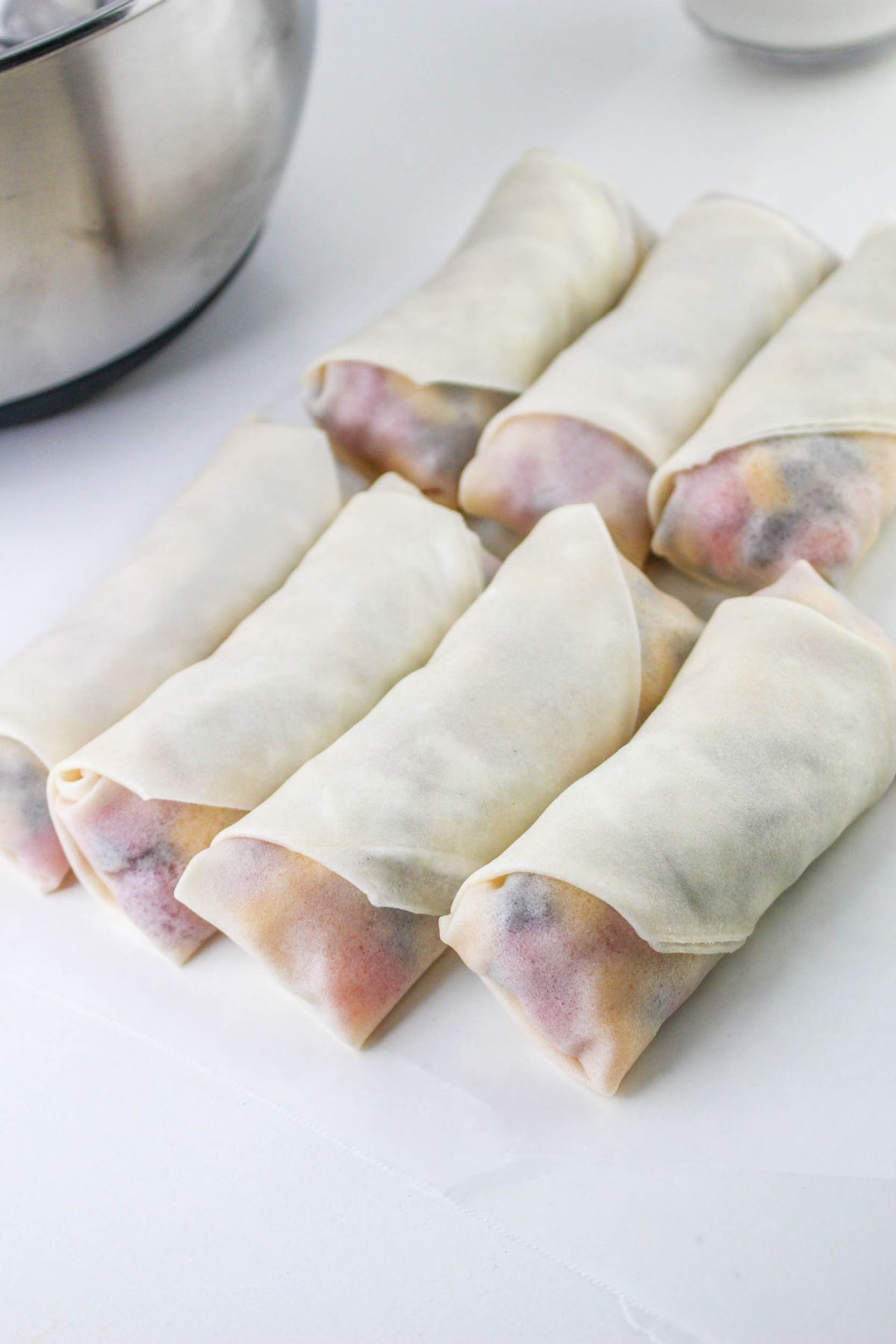 Eight uncooked spring rolls filled with assorted ingredients are arranged on a white surface, with a metal bowl visible in the background.