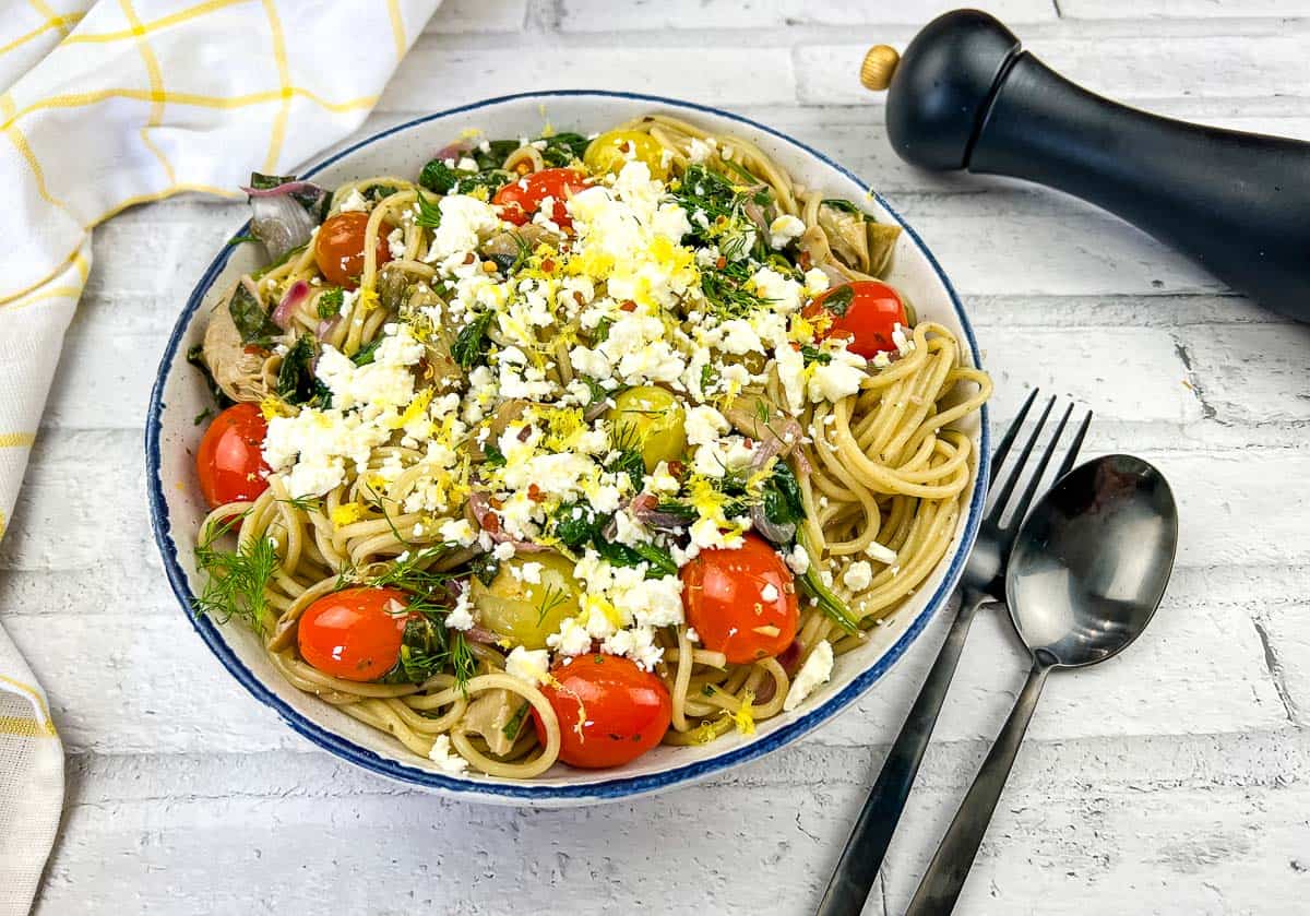 A bowl of spaghetti topped with cherry tomatoes, greens, shredded chicken, crumbled cheese, and herbs next to a fork, spoon, and black pepper grinder on a white surface.