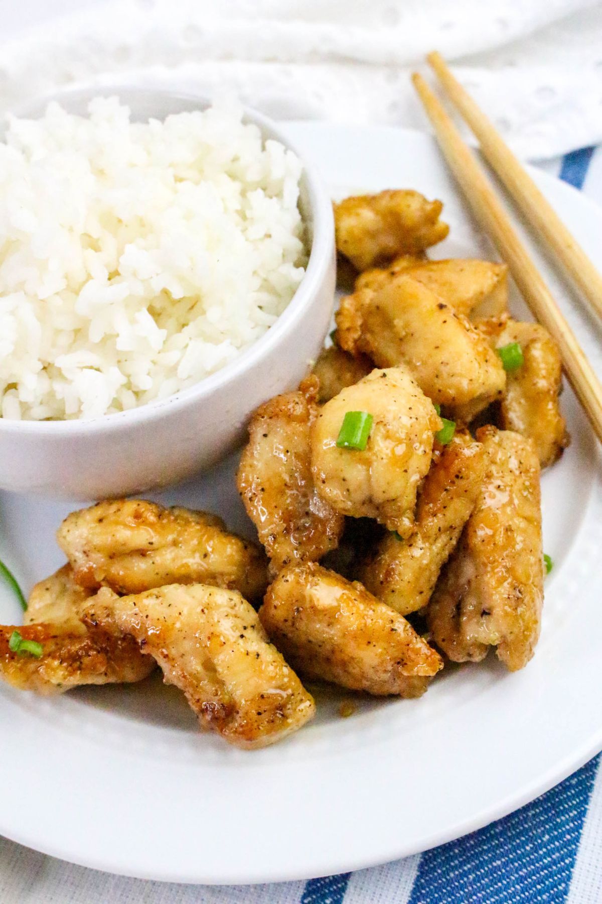 A plate with white rice in a small bowl and pieces of cooked chicken garnished with green onions, accompanied by chopsticks.