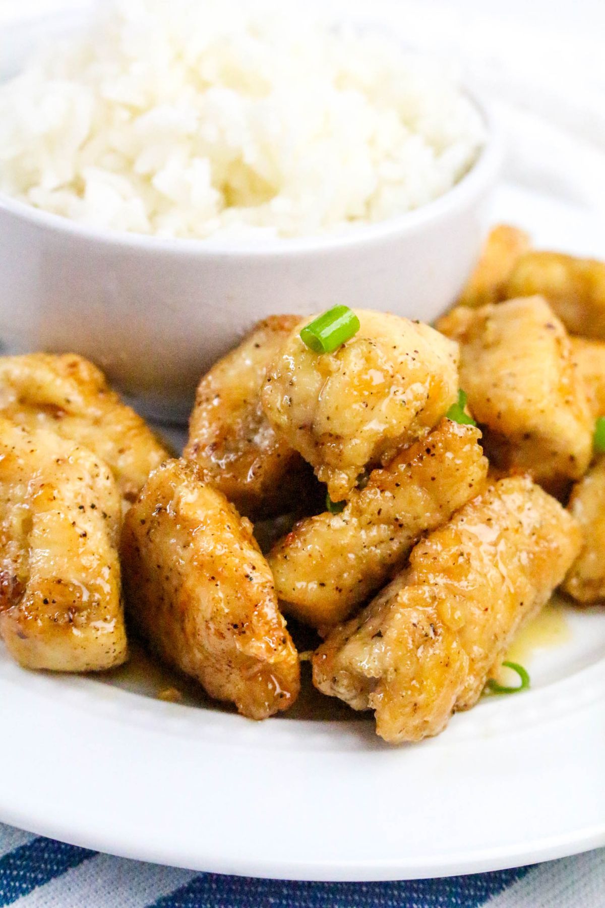 A plate of glazed, seasoned chicken pieces garnished with chopped green onions, served with a bowl of white rice in the background.