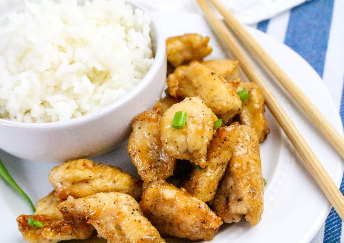 A plate with pieces of seasoned, cooked chicken next to a bowl of white rice, garnished with green onions and accompanied by wooden chopsticks.