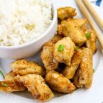 A plate of white rice served in a bowl alongside pieces of seasoned, cooked chicken garnished with green onions; chopsticks rest on the plate.