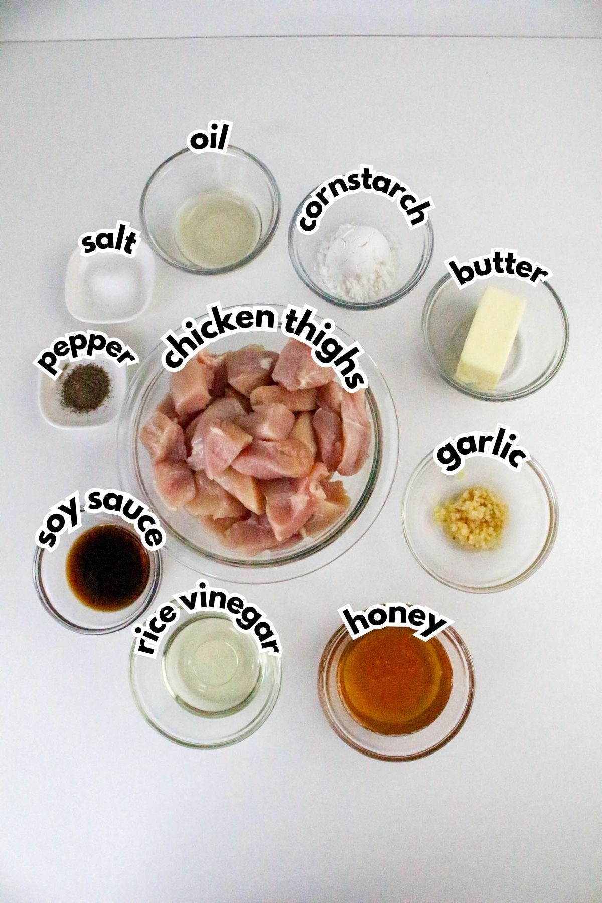 Top-down view of labeled ingredients in bowls for a chicken dish, including chicken thighs, oil, cornstarch, butter, salt, pepper, soy sauce, rice vinegar, garlic, and honey.