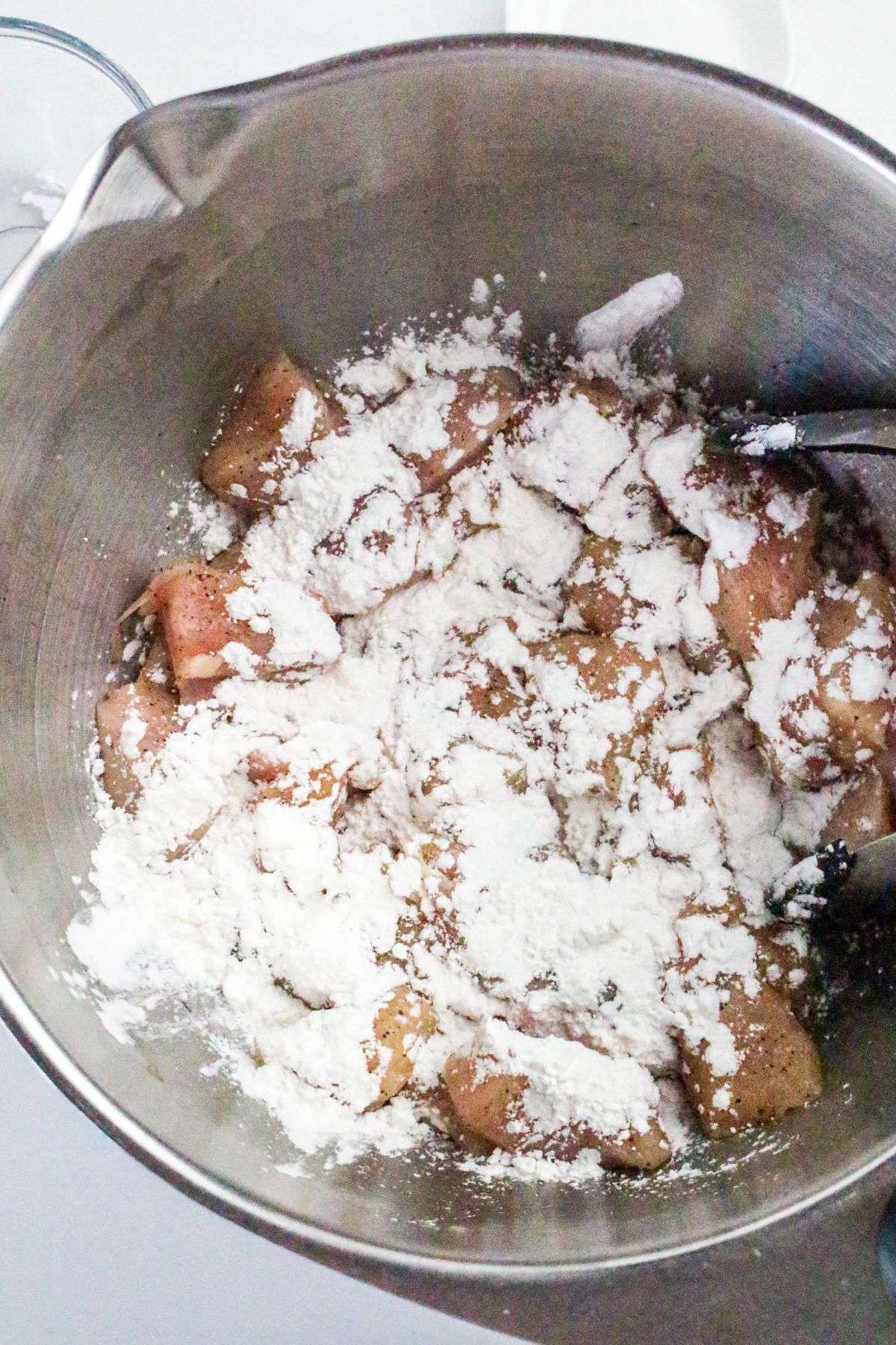 Metal mixing bowl with diced chicken pieces coated in a layer of white flour, with a spoon partially visible on the right side.