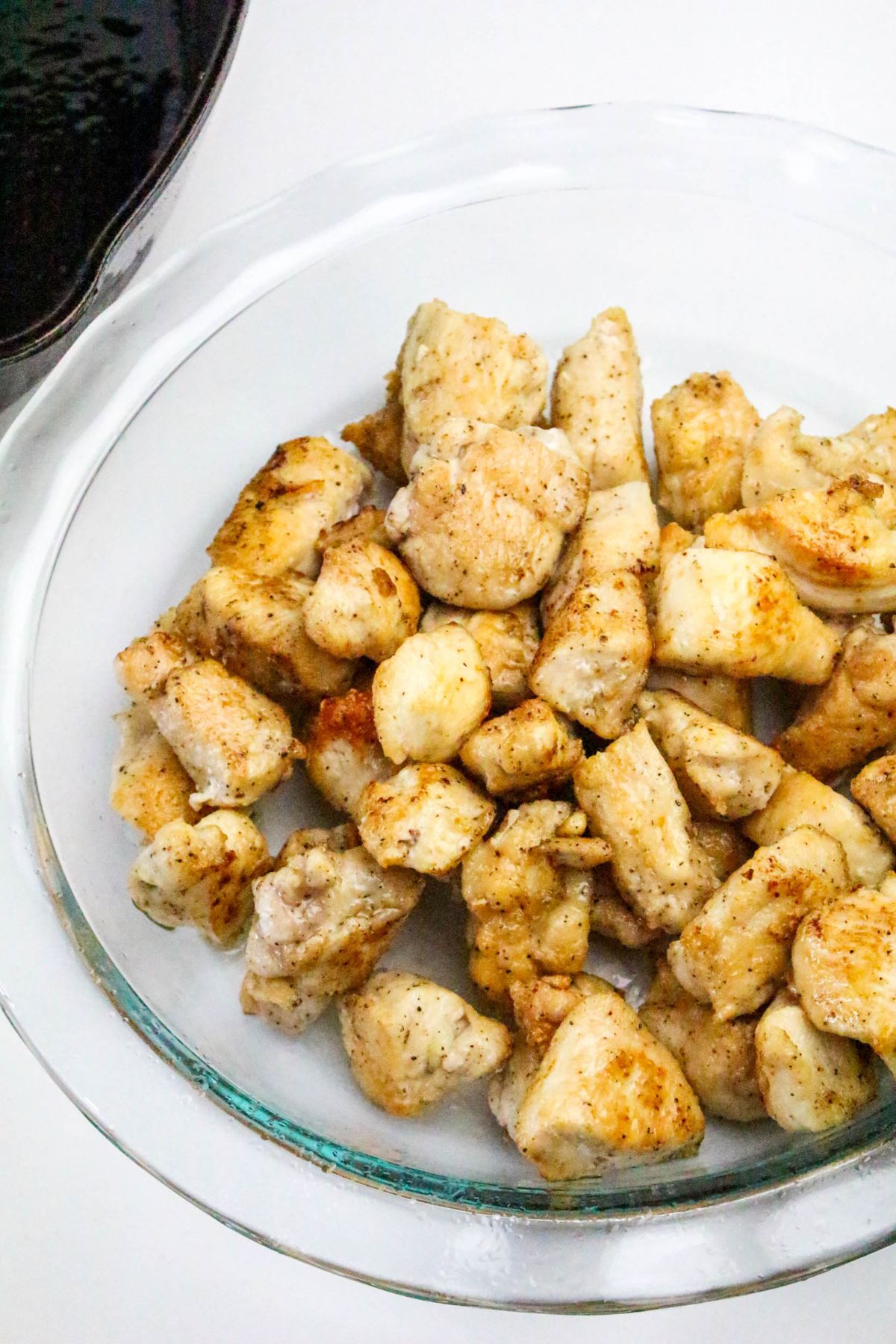 A clear glass dish filled with cooked, seasoned chicken breast pieces sits on a white surface near a black pan.