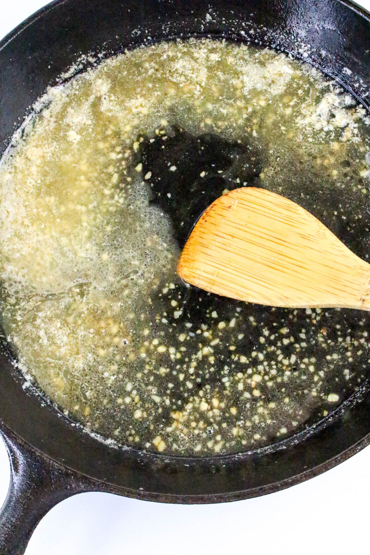 Minced garlic is sautéing in melted butter in a black cast iron skillet, stirred with a wooden spatula.