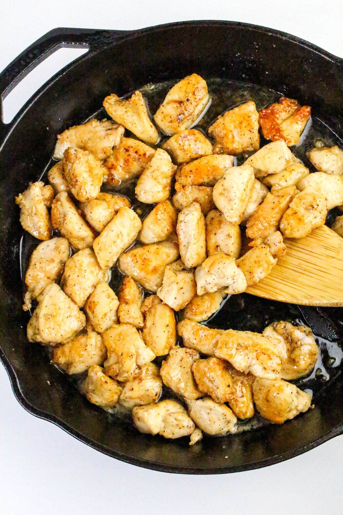 Cubed pieces of chicken breast cooking in a black cast iron skillet, being stirred with a wooden spatula.