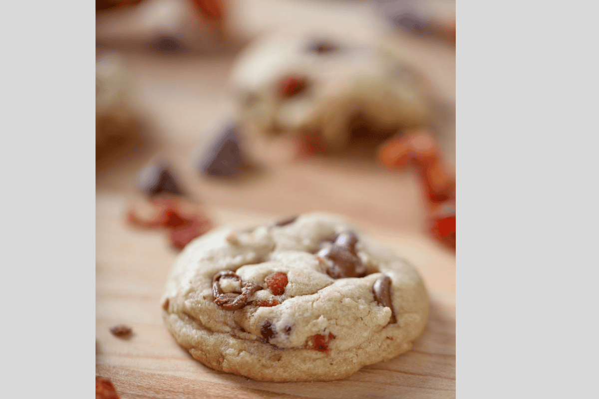 A close-up of a chocolate chip cookie with visible chocolate chunks and possibly caramel bits on a wooden surface, with blurred cookies and ingredients in the background.