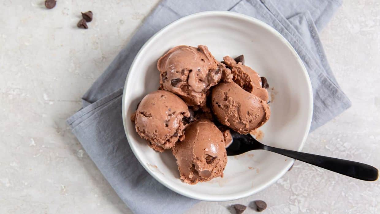 A white bowl with four scoops of chocolate ice cream and chocolate chips, placed on a gray napkin with a black spoon.
