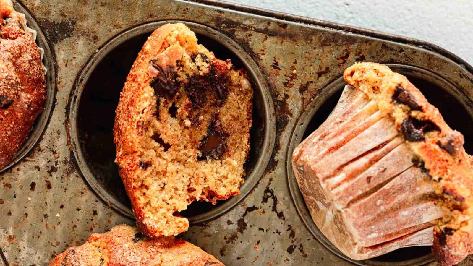 Three chocolate chip muffins in a metal muffin tin, with one muffin cut in half to show the chocolate chips inside.