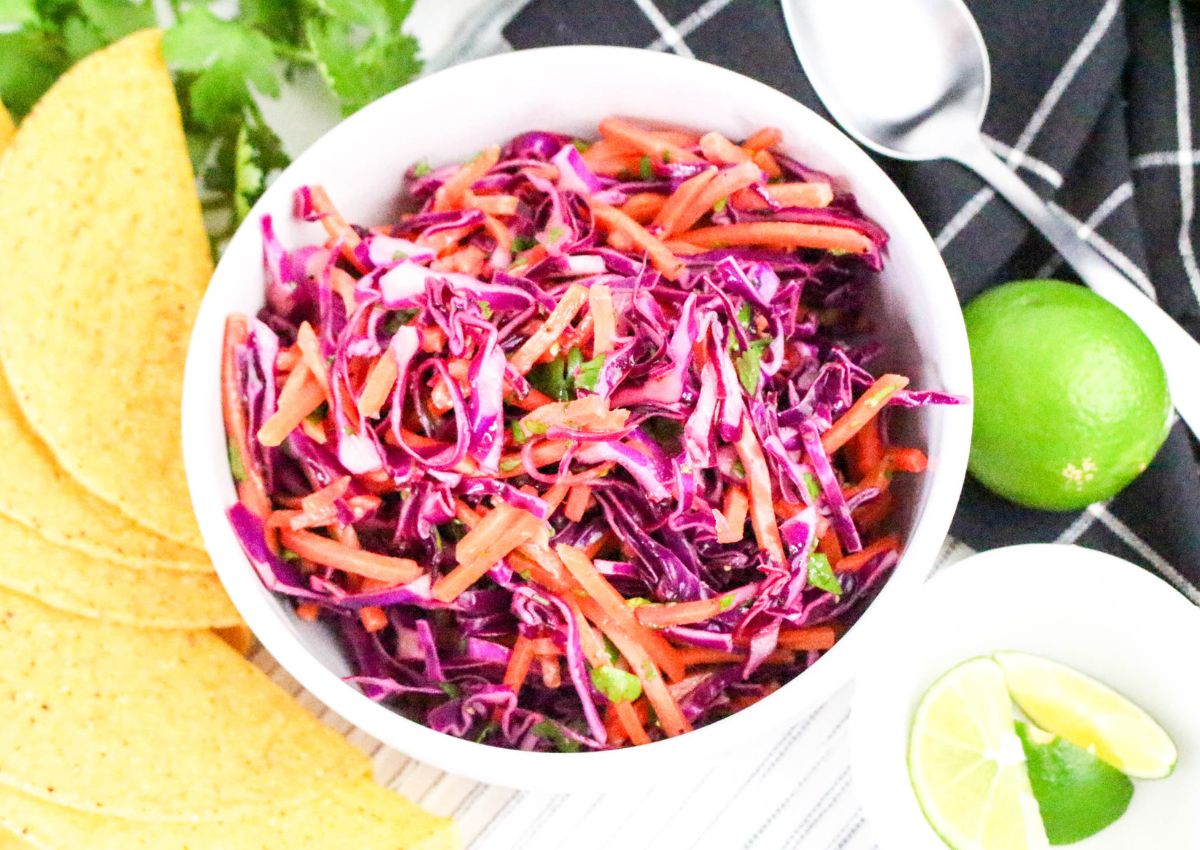 A white bowl filled with shredded red cabbage, carrots, and cilantro sits on a table next to taco shells, a lime, lime wedges, a spoon, and a black napkin.