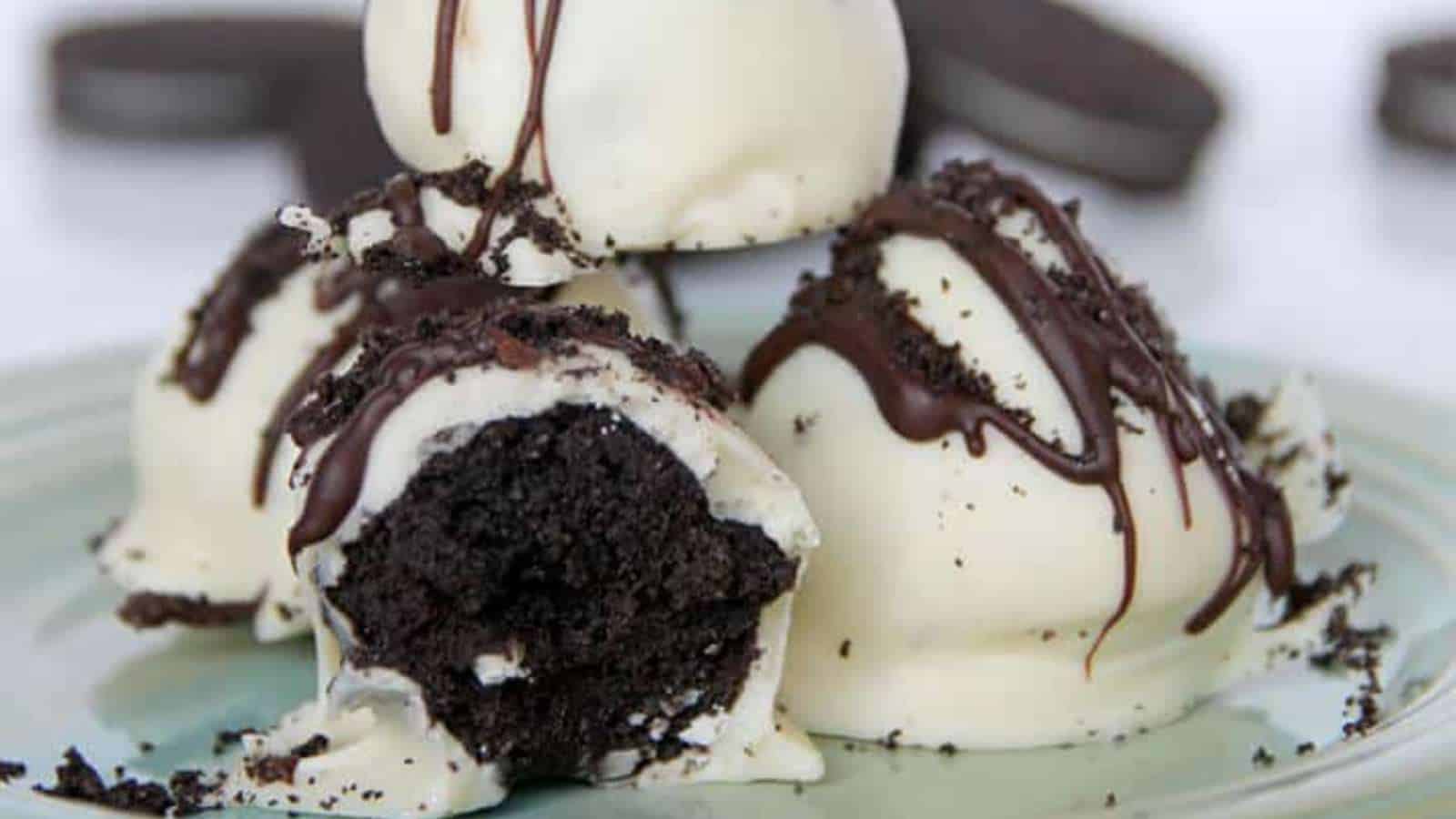 Three Oreo truffles coated in white chocolate, drizzled with dark chocolate, and sprinkled with crushed cookies. One truffle is partially bitten to show the dark cookie filling.