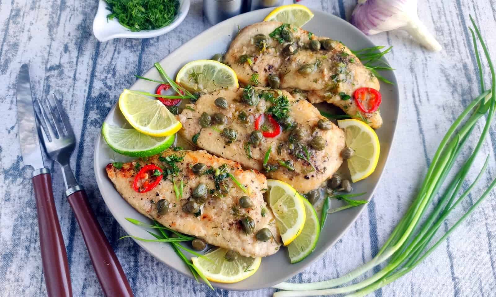 Three cooked chicken breasts garnished with capers, lemon slices, red chili, and fresh herbs on a plate, with forks, dill, green onions, and garlic nearby.