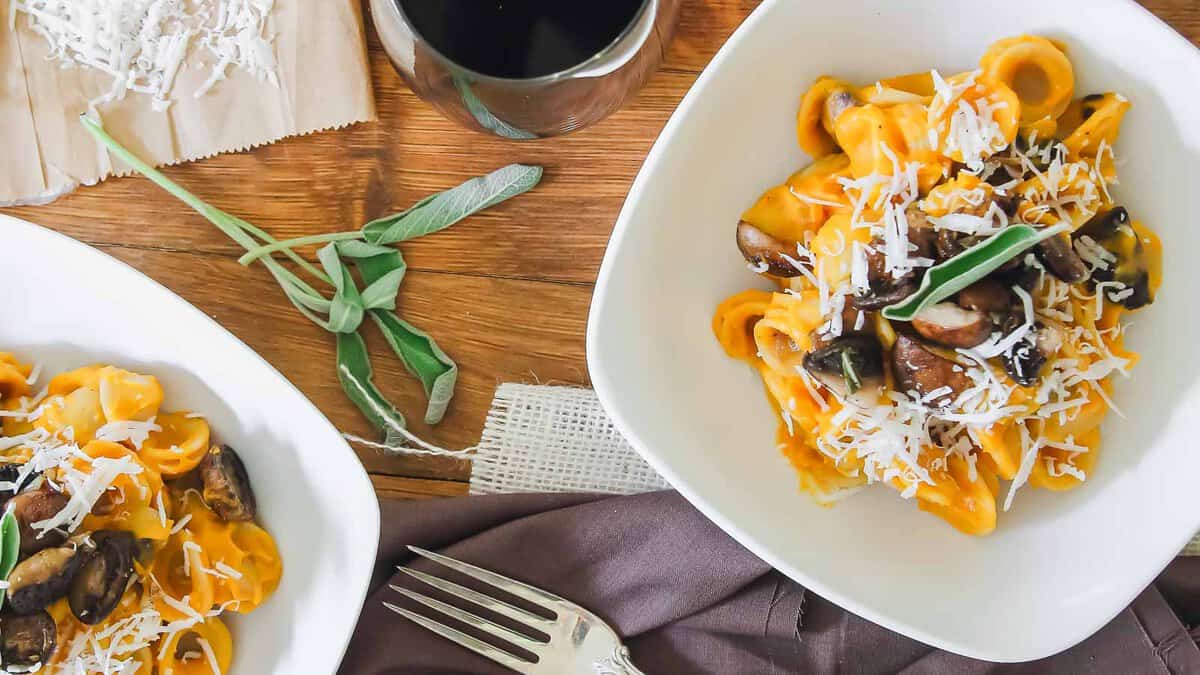 Two white bowls of pasta topped with mushrooms, grated cheese, and fresh sage sit on a wooden table next to a fork, napkin, shredded cheese, and a glass of red wine.