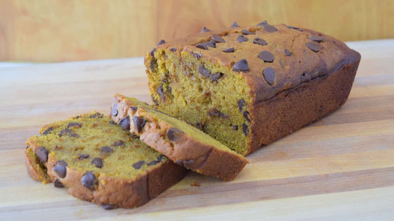 A loaf of pumpkin bread with chocolate chips sits on a wooden surface, with two slices cut and laid in front.