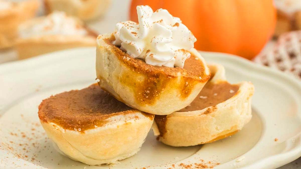 Three mini pumpkin pies are stacked on a plate, with one topped with whipped cream and a sprinkle of cinnamon.