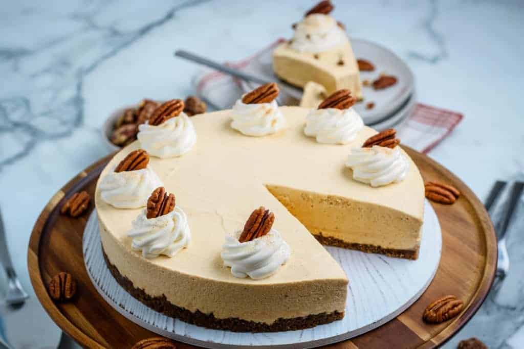 A whole cheesecake with a sliced piece on a plate, topped with whipped cream and pecans, sits on a wooden platter with pecans scattered around.