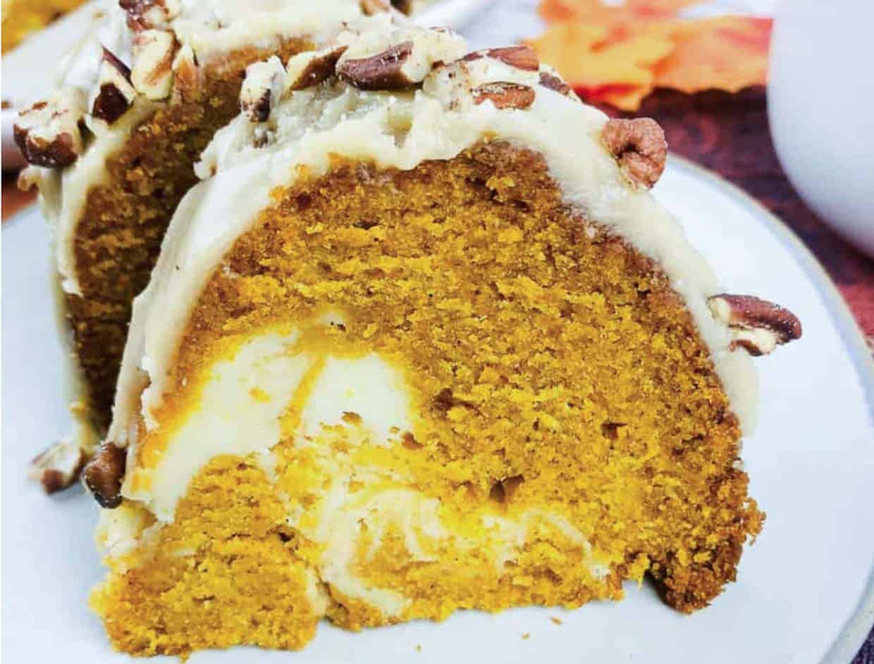 A slice of pumpkin Bundt cake with a cream cheese swirl, topped with frosting and pecans, on a white plate.