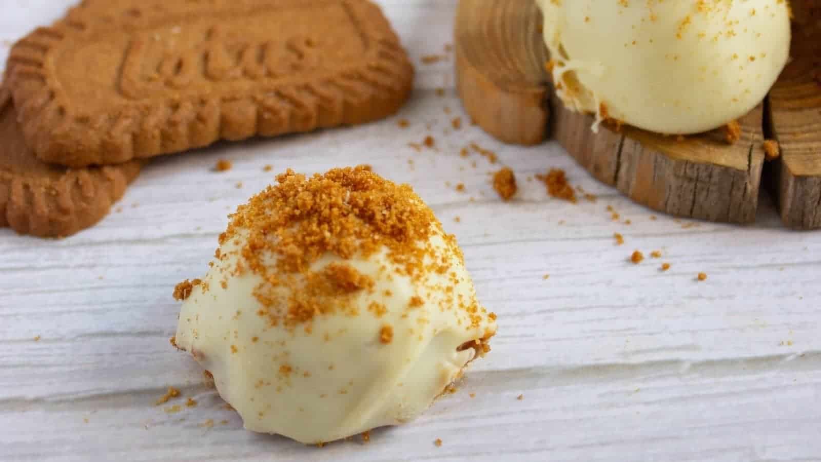 A white chocolate truffle topped with cookie crumbs sits on a white surface, with two Lotus Biscoff cookies and another truffle in the background.