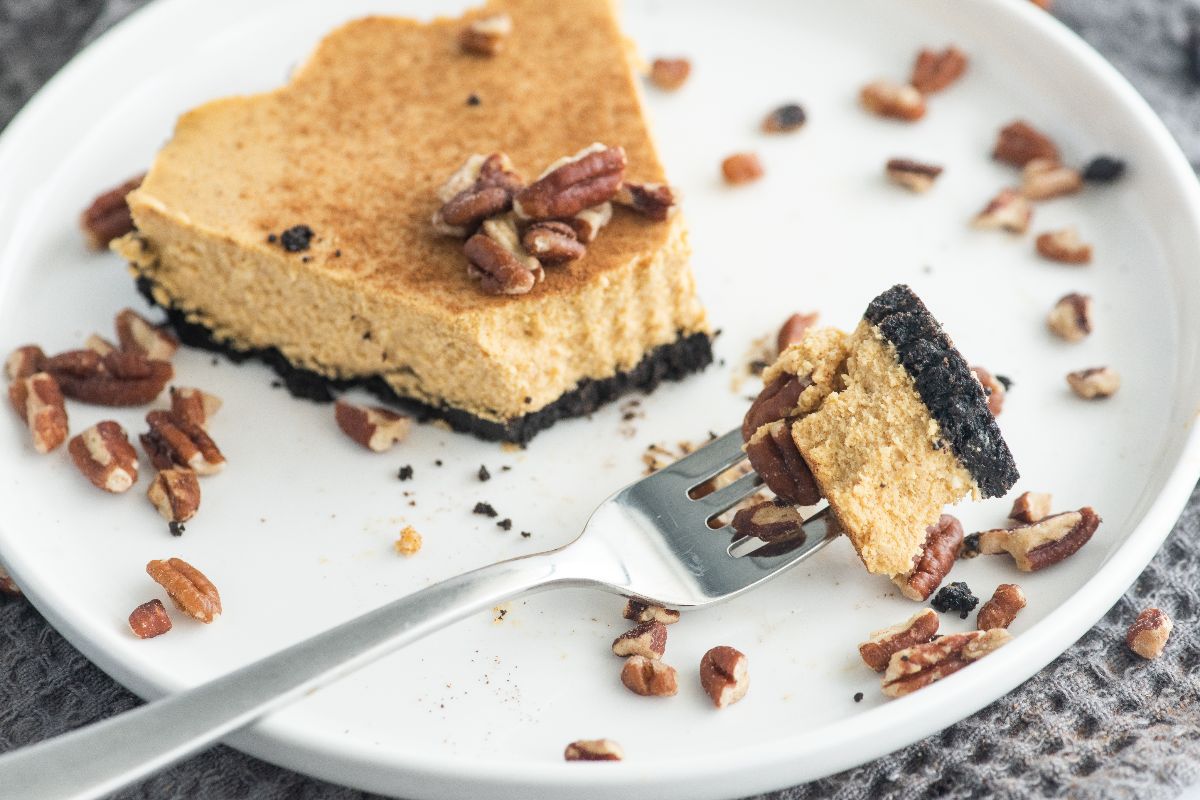A partially eaten slice of pumpkin cheesecake with a dark cookie crust sits on a white plate, topped and surrounded by pecans; a fork holds a bite-sized piece.