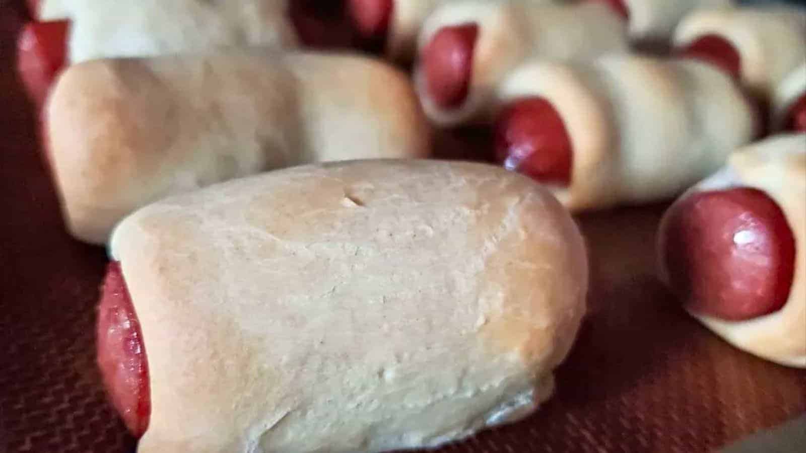 Close-up of several pigs in a blanket, showing small sausages wrapped in baked dough, arranged on a brown surface.