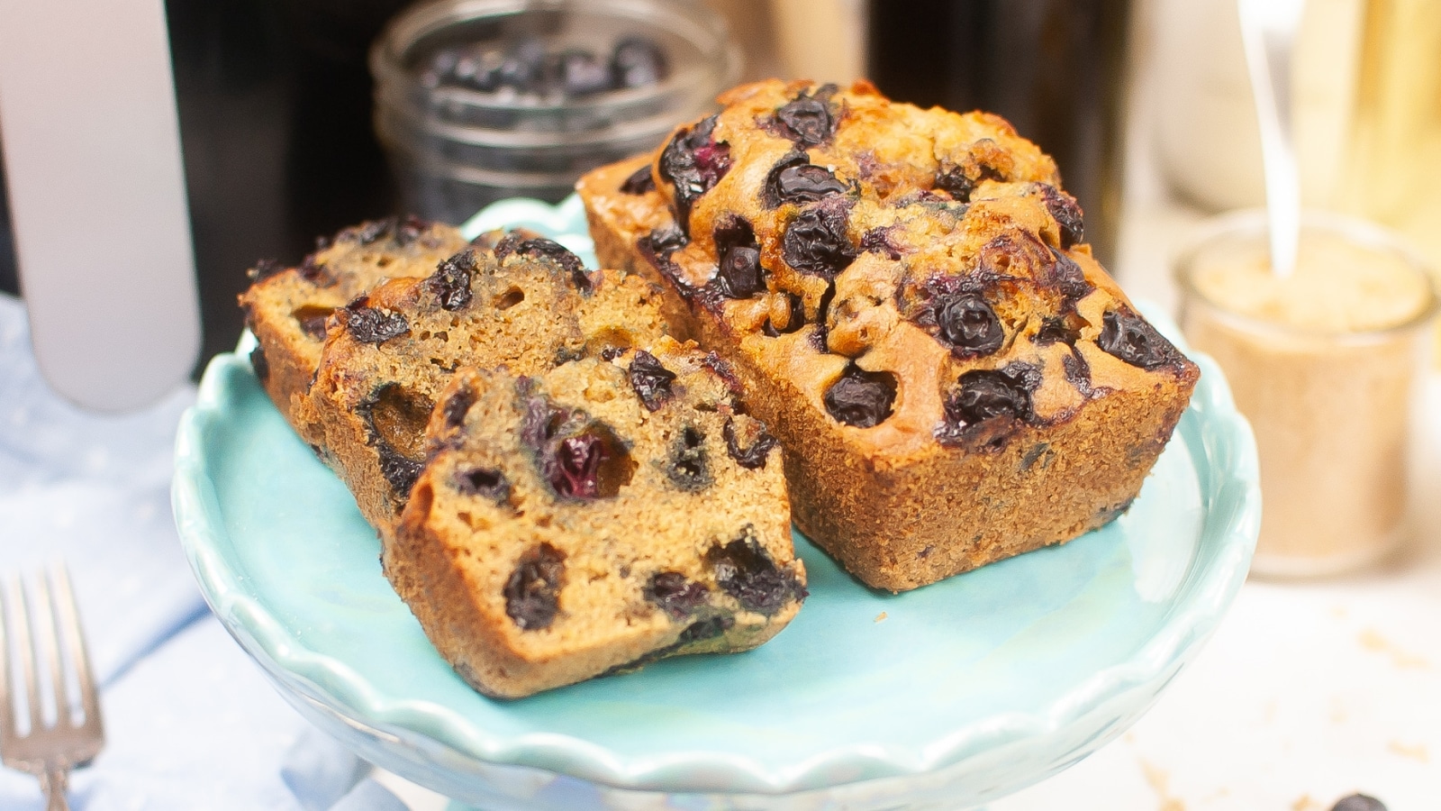 Two slices and one whole loaf of blueberry bread sit on a light blue cake stand, with more blueberries and baking ingredients in the background.