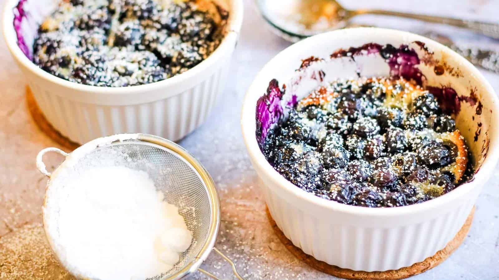 Two white ramekins filled with baked blueberry dessert, dusted with powdered sugar, sit on cork coasters next to a small sieve containing more powdered sugar.